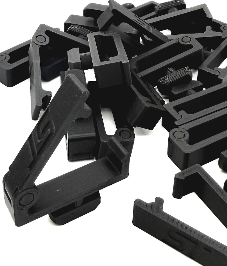 May include: A pile of black plastic clips, each with a rectangular frame and a hinged arm. The clips have a matte finish and are designed to connect or secure items. The image is taken against a white background.