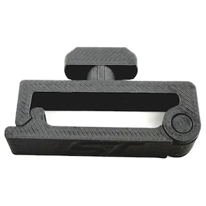 May include: A black plastic buckle with a rectangular frame and a central handle. The buckle has a textured surface and a circular element on one end. The handle is approximately 1 inch long.
