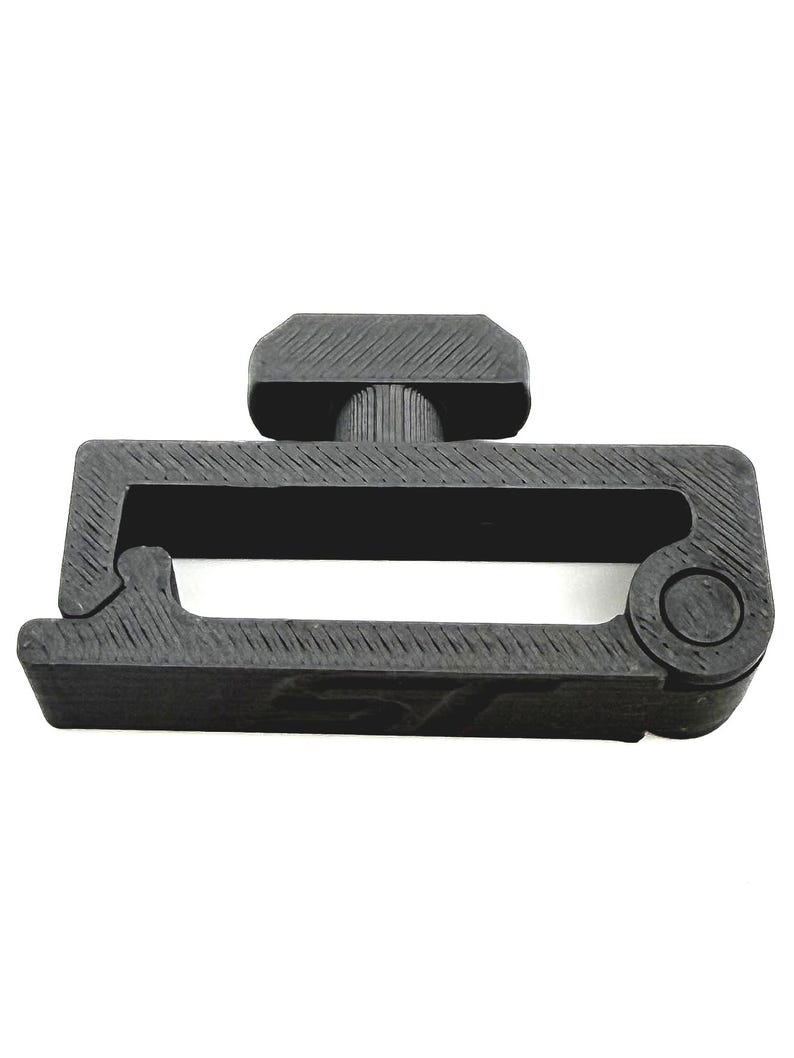 May include: A black plastic buckle with a rectangular frame and a central handle. The buckle has a textured surface and a circular element on one end. The handle is approximately 1 inch long.