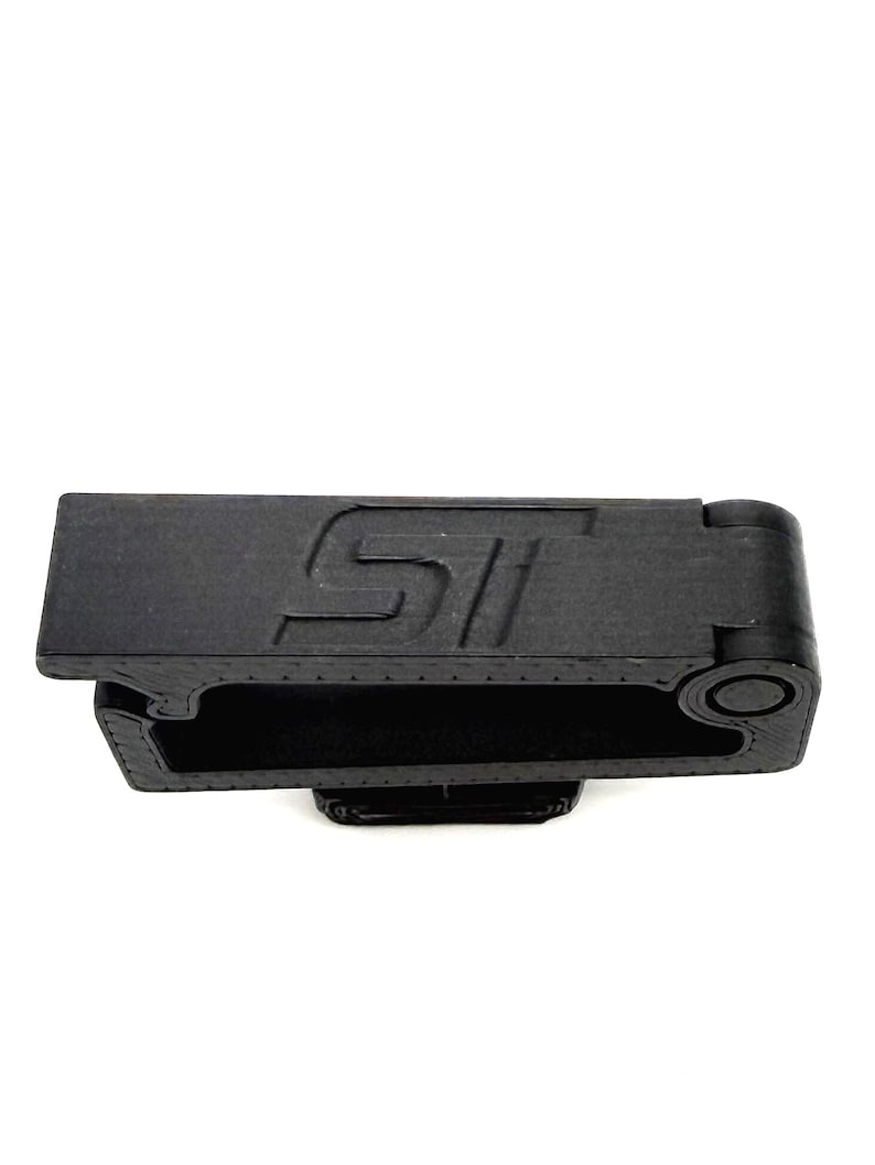 May include: A black, rectangular object with the letters "ST" embossed on the top surface. The object has a hollow interior and a rounded end with a circular detail. The item is made of a matte material.