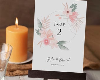 May include: A wedding table number card with floral accents. The card displays "TABLE 2" and the names "Julia & Daniel" with a date. The card is held by a small wooden easel, with a lit candle and a glass in the background.