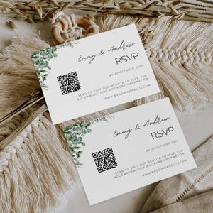May include: Two RSVP cards with the names "Emmy & Andrew" and the text "RSVP" in elegant script. The cards feature a QR code and the website address "www.weddingwebsite.com". Greenery decorates the upper left corner of each card.