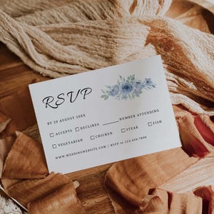May include: A white RSVP card with blue floral accents. The card includes fields for accepting or declining, meal choices, and the number attending. The card is on a wooden surface with brown fabric.