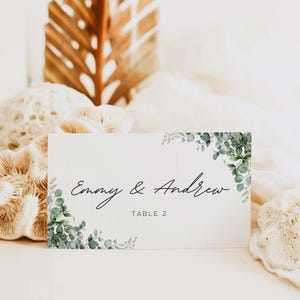 Puede incluir: Una tarjeta de mesa blanca con los nombres "Emmy & Andrew" en escritura cursiva y "TABLE 2" debajo. Acentos de vegetación verde en las esquinas. La tarjeta está sobre un fondo suave y neutro, ideal para bodas o eventos.