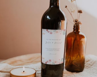 Puede incluir: Una botella de vino oscuro con una etiqueta personalizada para una boda, con detalles florales y los nombres "Jasen & Daniel". También hay una pequeña vela dorada y un jarrón de vidrio marrón con flores secas.