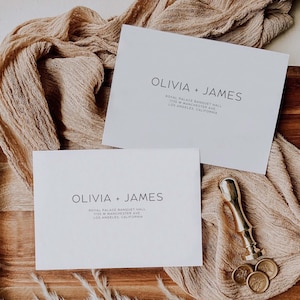 Puede incluir: Dos tarjetas de invitación blancas con el texto "OLIVIA + JAMES" y detalles de la dirección. Un sello de cera dorado y tres sellos de cera están sobre una superficie de madera con un fondo de tela beige. Papelería de boda.