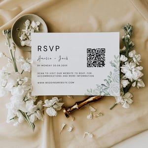 Puede incluir: Una tarjeta RSVP blanca con detalles florales y un código QR, rodeada de flores blancas y un sello de cera dorado. La tarjeta incluye los nombres "Amelia + Jack" e información del sitio web.