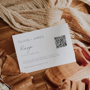 May include: A white RSVP card with the names Olivia and James, a QR code, and wedding website details. The card rests on a wooden surface with tan fabric and ribbons.