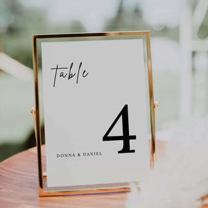 May include: A gold-framed table number card with the word "table" in script and the number "4" in a bold font. The names "DONNA & DANIEL" are printed below the number. The card is displayed on a wooden surface.