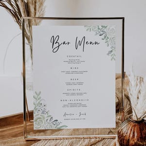 Könnte beinhalten: Ein gerahmtes Bar-Menü mit floralen Verzierungen. Die Speisekarte listet Cocktails, Wein, Bier, Spirituosen und alkoholfreie Getränke auf. Der Text enthält die Namen der Getränke, die Namen Amelia & Jack und das Datum.