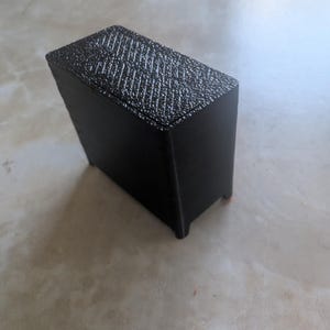 Puede incluir: Una caja rectangular negra con una superficie superior texturizada. La caja tiene un acabado mate y pequeños pies. La superficie superior tiene un patrón entrecruzado. La caja probablemente está impresa en 3D.