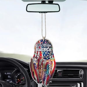 Peut inclure: Un désodorisant de voiture avec un design patriotique. Le haut rond porte le texte "God Bless AMERICA" sur un fond bleu avec des étoiles blanches. Le bas a un design à rayures rouges et blanches avec des plumes colorées.