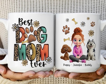 May include: Two white ceramic mugs with handles. One mug reads "Best Dog Mom Ever" with paw prints and floral accents. The other mug features a cartoon of a girl with two dogs and the text "Puppy - Jennifer - Buddy".