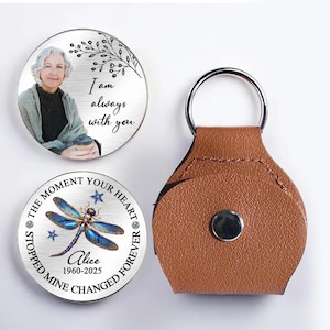 Pode incluir: Um conjunto de chaveiros comemorativos. Um chaveiro apresenta uma foto de uma mulher e o texto "I am always with you". Outro chaveiro tem um design de libélula e o texto "The moment your heart stopped mine changed forever". Um porta-chaves de couro castanho também está incluído.