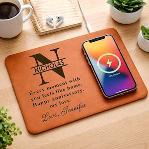 May include: A brown leather wireless charging pad with a phone charging on top. The pad has the name "Nicholas" and a message engraved on it. A coffee mug, notepad, and small plants are also visible.