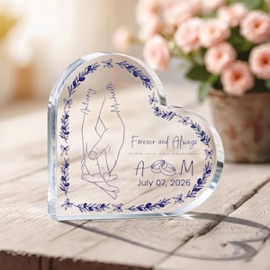 May include: A heart-shaped clear acrylic plaque featuring a blue printed design of two hands holding, a floral border, and the text: Antony, Michelle, Forever and Always, A M, and July 07, 2026. The plaque includes a graphic of two interlocking rings.