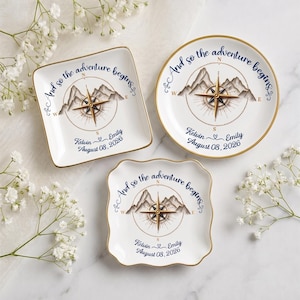 May include: Three white ceramic dishes with gold rims, featuring a compass and mountain illustration, the text "And so the adventure begins," and the names "Kelvin & Emily" with the date "August 08, 2026" printed in dark blue ink.