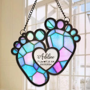 May include: A stained glass baby feet ornament with a heart in the center. The heart has the name Adeline, 7 lbs, 7:15 AM, 23.03.2024 inscribed on it. The feet are blue and pink with a mosaic pattern.