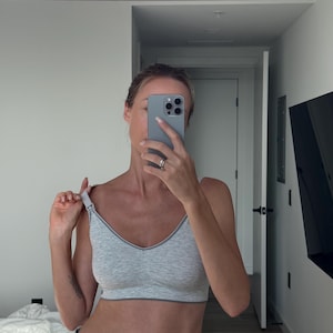 May include: A light gray sports bra with a V-neck and a thin gray trim. The bra is made of a soft, stretchy material. The person is wearing blue denim jeans.