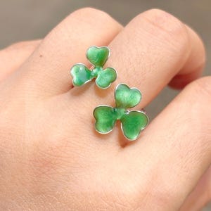 May include: Two silver rings, each featuring a three-leaf clover design with bright green enamel. The clovers have a shiny finish and are set against a neutral background. The rings are a unique and eye-catching accessory.
