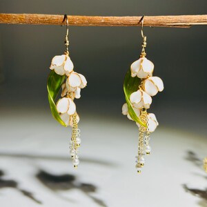 Op de afbeelding: Een paar handgemaakte oorbellen met witte lelietjes-van-dalen bloemen, gouden accenten en bungelende parels. De oorbellen bevatten groene bladeren en goudkleurige haken, wat een elegant bloemendesign cre&euml;ert.
