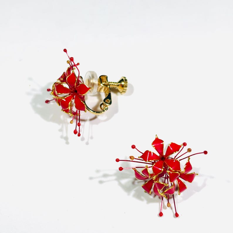 K&ouml;nnte beinhalten: Ein Paar Clip-Ohrringe mit floralem Design. Die Ohrringe sind &uuml;berwiegend rot mit goldenen Akzenten und feinen Drahtdetails. Der Clip-Mechanismus ist goldfarben.