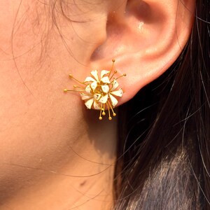K&ouml;nnte beinhalten: Nahaufnahme eines wei&szlig;en und goldenen floralen Ohrsteckers. Der Ohrring hat eine Ansammlung wei&szlig;er Bl&uuml;tenbl&auml;tter mit goldenen Akzenten und filigranen Golddrahtdetails. Der Ohrring wird an einem Ohr getragen.
