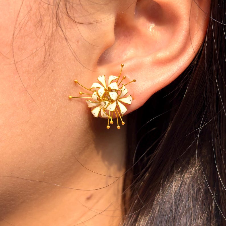 K&ouml;nnte beinhalten: Nahaufnahme eines wei&szlig;en und goldenen floralen Ohrsteckers. Der Ohrring hat eine Ansammlung wei&szlig;er Bl&uuml;tenbl&auml;tter mit goldenen Akzenten und filigranen Golddrahtdetails. Der Ohrring wird an einem Ohr getragen.