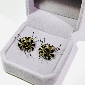 K&ouml;nnte beinhalten: Ein Paar schwarze und goldfarbene Blumenohrringe in einer grauen Samt-Schmuckschatulle. Die Ohrringe haben ein einzigartiges Design mit schwarzen Bl&uuml;tenbl&auml;ttern, goldenen Akzenten und filigranen schwarzen Drahtdetails.