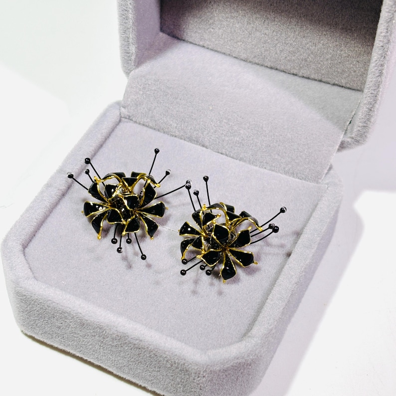 K&ouml;nnte beinhalten: Ein Paar schwarze und goldfarbene Blumenohrringe in einer grauen Samt-Schmuckschatulle. Die Ohrringe haben ein einzigartiges Design mit schwarzen Bl&uuml;tenbl&auml;ttern, goldenen Akzenten und filigranen schwarzen Drahtdetails.