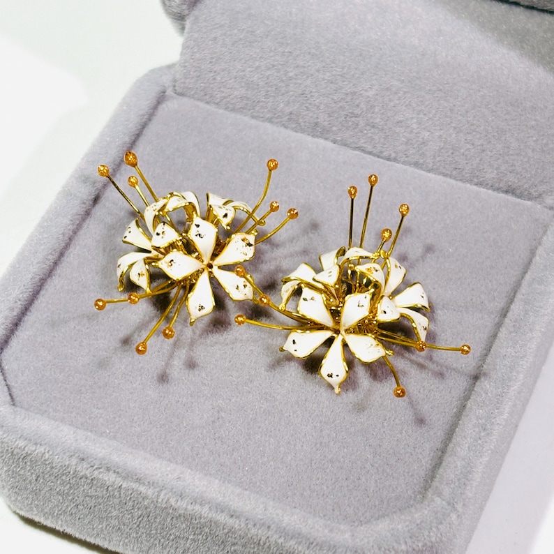 K&ouml;nnte beinhalten: Ein Paar goldfarbene Blumenohrringe mit wei&szlig;en Emaille-Bl&uuml;tenbl&auml;ttern und filigranen Goldakzenten. Die Ohrringe werden in einer grauen Samt-Schmuckschatulle pr&auml;sentiert, die ihr aufwendiges Design hervorhebt. Ein einzigartiges Accessoire.