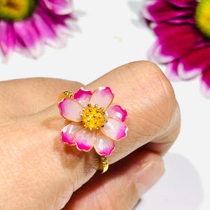 May include: A gold ring featuring a pink and white flower design with a yellow center. The flower petals have a bright pink outline. The ring is set against a soft background with hints of pink flowers.