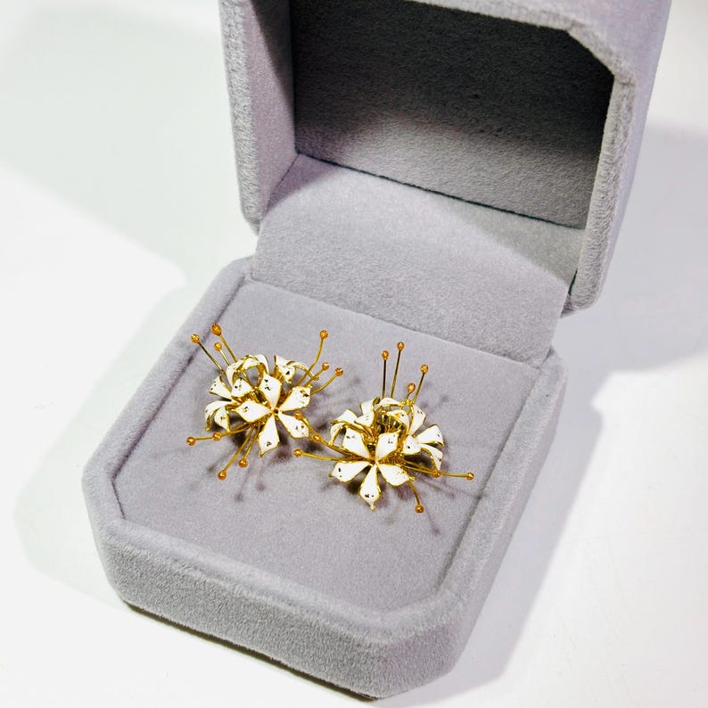 K&ouml;nnte beinhalten: Ein Paar wei&szlig;e und goldfarbene Blumenohrringe, pr&auml;sentiert in einer grauen Samt-Schmuckschatulle. Die Ohrringe haben ein Golddraht-Design mit wei&szlig;en Emaille-Bl&uuml;tenbl&auml;ttern und kleinen goldenen Akzenten. Die Schatulle ist ge&ouml;ffnet und zeigt die Ohrringe.
