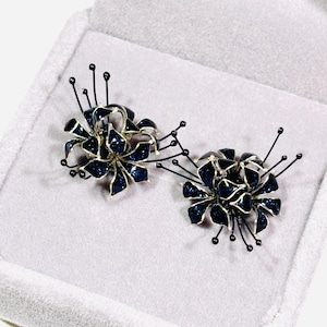K&ouml;nnte beinhalten: Ein Paar bl&uuml;tenf&ouml;rmige Ohrringe. Jeder Ohrring hat ein dunkelblaues, glitzerndes Blumendesign mit silberfarbenen Umrissen. Schwarze Stiele mit kleinen schwarzen Perlen erstrecken sich von den Bl&uuml;ten. Die Ohrringe werden in einer hellgrauen Schmuckschatulle pr&auml;sentiert.