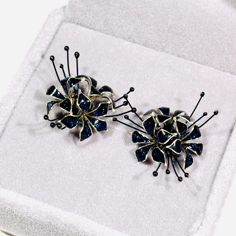 K&ouml;nnte beinhalten: Ein Paar bl&uuml;tenf&ouml;rmige Ohrringe. Jeder Ohrring hat ein dunkelblaues, glitzerndes Blumendesign mit silberfarbenen Umrissen. Schwarze Stiele mit kleinen schwarzen Perlen erstrecken sich von den Bl&uuml;ten. Die Ohrringe werden in einer hellgrauen Schmuckschatulle pr&auml;sentiert.
