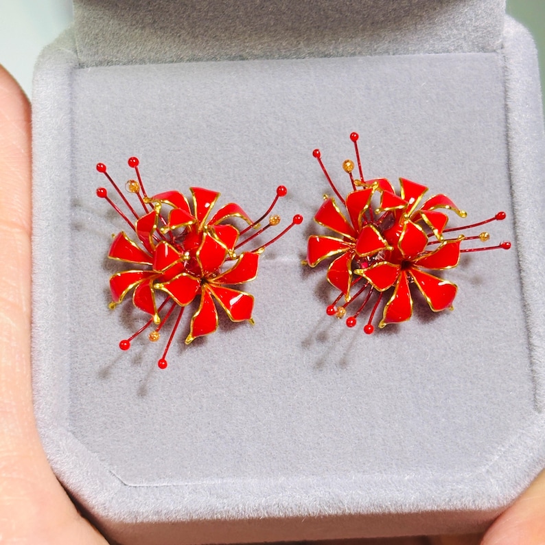 K&ouml;nnte beinhalten: Ein Paar rot-goldene, bl&uuml;tenf&ouml;rmige Ohrringe. Jeder Ohrring hat eine Ansammlung roter Bl&uuml;tenbl&auml;tter mit goldenen Verzierungen und zarten roten und goldenen Drahtakzenten. Die Ohrringe werden in einer grauen Schmuckschatulle pr&auml;sentiert.