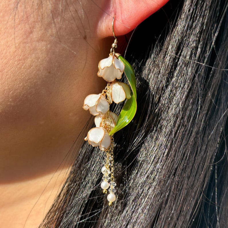 Op de afbeelding: Enkele goudkleurige oorbel met witte bloesems met gouden randen, een groen blad en bungelende imitatieparels. Het ontwerp lijkt op lelietjes-van-dalen bloemen.