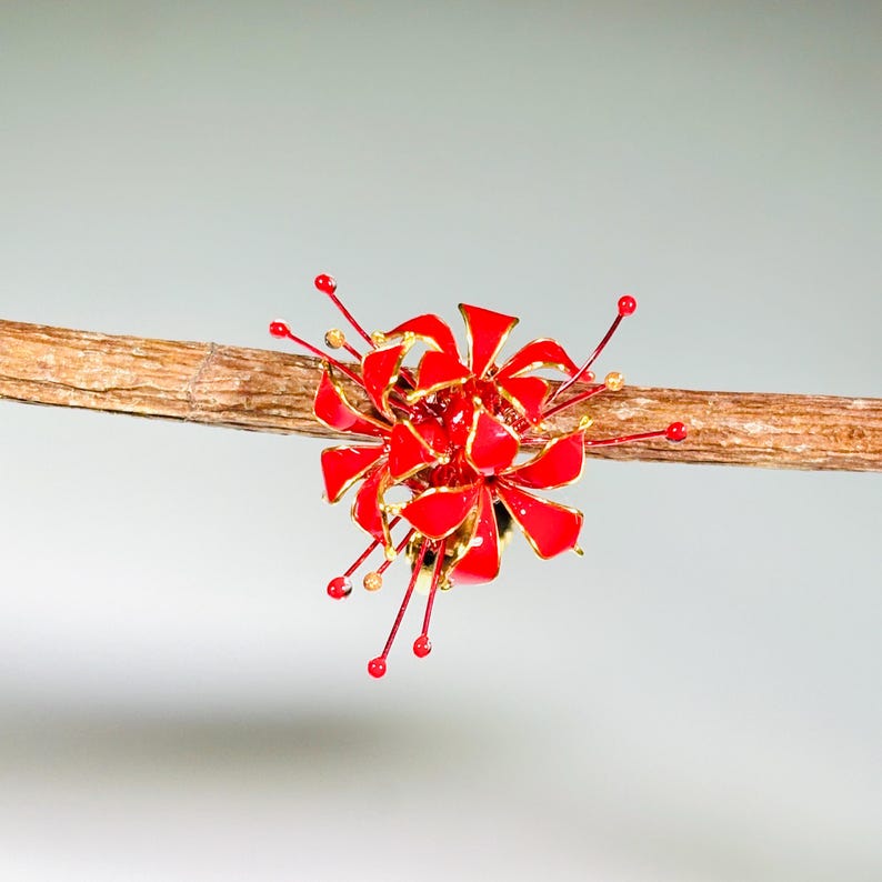 K&ouml;nnte beinhalten: Ein leuchtend rotes und goldenes Blumenornament, m&ouml;glicherweise eine Brosche oder ein Haaraccessoire, sitzt auf einem schlanken, braunen Ast. Das Ornament hat mehrere Bl&uuml;tenbl&auml;tter und zarte, l&auml;ngliche Akzente, die einen auff&auml;lligen Kontrast erzeugen.