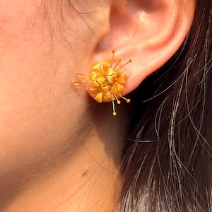 K&ouml;nnte beinhalten: Nahaufnahme eines goldfarbenen floralen Ohrsteckers. Der Ohrring hat eine Ansammlung von Bl&uuml;tenbl&auml;ttern und zarten, strahlenf&ouml;rmigen Spitzen, die ein dreidimensionales Design bilden. Der Ohrring wird an einem Ohr getragen und zeigt seine komplizierten Details und das helle, metallische Finish.