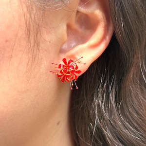 K&ouml;nnte beinhalten: Nahaufnahme eines roten und goldenen Blumenohrsteckers. Der Ohrring hat eine Ansammlung roter Bl&uuml;tenbl&auml;tter mit goldenen Akzenten und kleinen goldenen und roten Details. Der Ohrring wird an einem Ohr getragen.