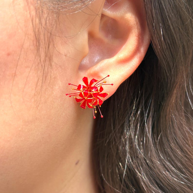 K&ouml;nnte beinhalten: Nahaufnahme eines roten und goldenen Blumenohrsteckers. Der Ohrring hat eine Ansammlung roter Bl&uuml;tenbl&auml;tter mit goldenen Akzenten und kleinen goldenen und roten Details. Der Ohrring wird an einem Ohr getragen.