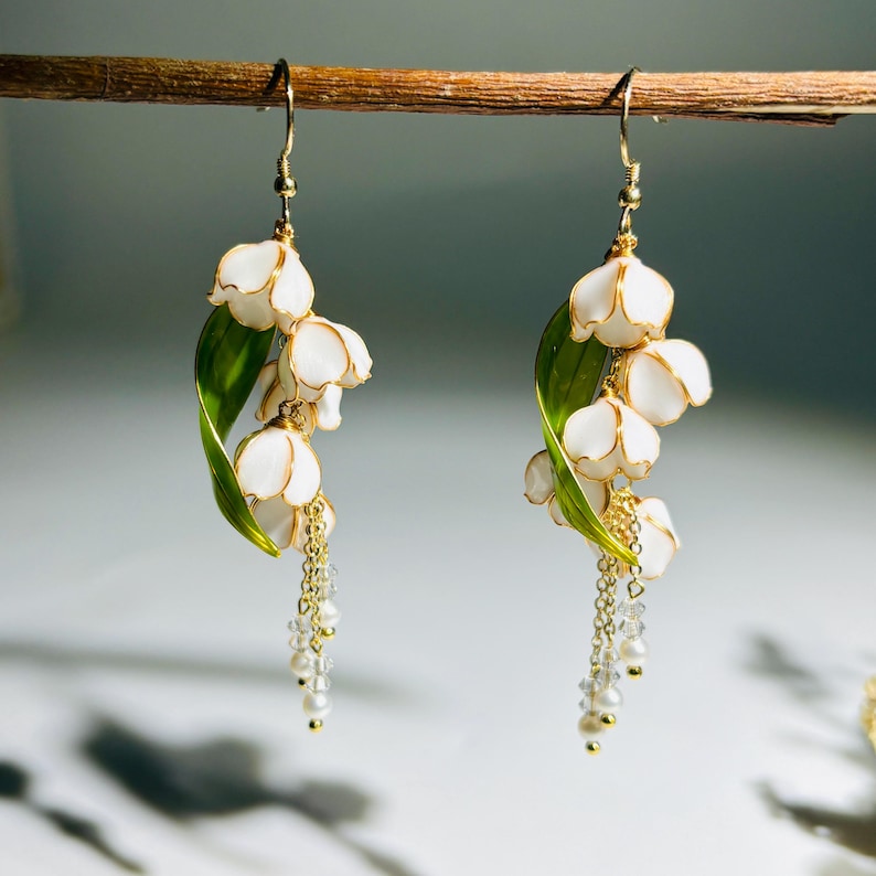 Op de afbeelding: Een paar witte bloemvormige oorbellen met gouden accenten en bungelende parels en kristallen kralen. De oorbellen hebben een lelietje-van-dalen-ontwerp met groene bladeren en goudkleurige haken. De oorbellen hangen aan een houten staaf.