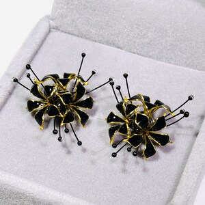 K&ouml;nnte beinhalten: Ein Paar schwarze und goldfarbene Blumenohrringe. Jeder Ohrring hat eine Ansammlung schwarzer Bl&uuml;tenbl&auml;tter mit goldenen Akzenten und zarten schwarzen Stielen mit kleinen, runden Verzierungen. Die Ohrringe werden in einer grauen Schmuckschatulle pr&auml;sentiert.