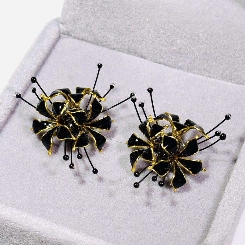 K&ouml;nnte beinhalten: Ein Paar schwarze und goldfarbene Blumenohrringe. Jeder Ohrring hat eine Ansammlung schwarzer Bl&uuml;tenbl&auml;tter mit goldenen Akzenten und zarten schwarzen Stielen mit kleinen, runden Verzierungen. Die Ohrringe werden in einer grauen Schmuckschatulle pr&auml;sentiert.
