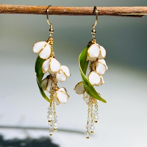 Op de afbeelding: Hangende oorbellen met witte bloemclusters, gouden accenten en groene bladeren. Elke oorbel heeft een delicate ketting met kleine, heldere kralen. De oorbellen zijn opgehangen aan goudkleurige haken.
