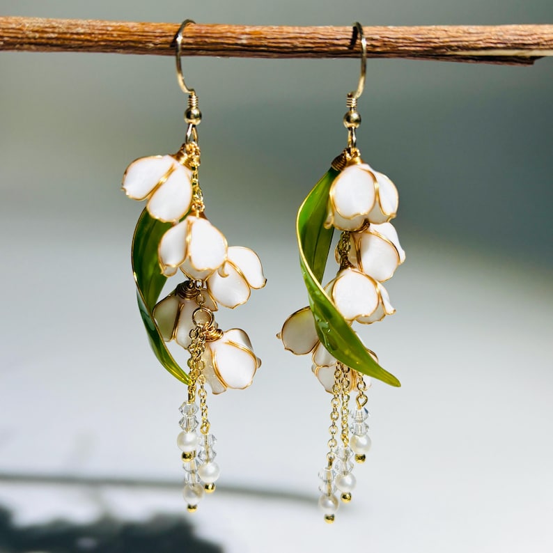 Op de afbeelding: Hangende oorbellen met witte bloemclusters, gouden accenten en groene bladeren. Elke oorbel heeft een delicate ketting met kleine, heldere kralen. De oorbellen zijn opgehangen aan goudkleurige haken.