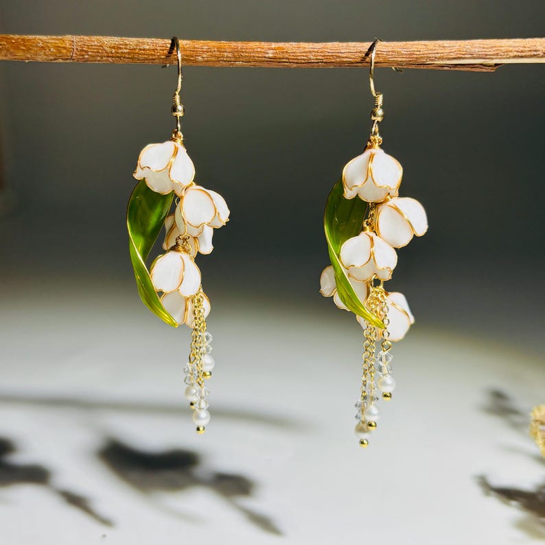 Op de afbeelding: Een paar handgemaakte oorbellen met witte lelietjes-van-dalen bloemen, gouden accenten en bungelende parels. De oorbellen bevatten groene bladeren en goudkleurige haken, wat een elegant bloemendesign cre&euml;ert.