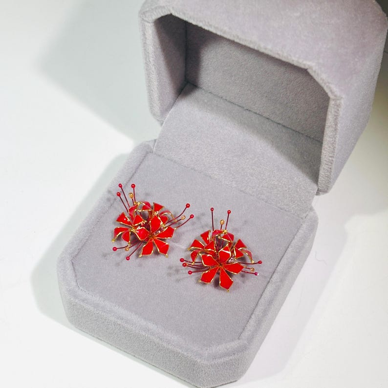 May include: A pair of red and gold flower-shaped earrings displayed in a gray velvet jewelry box. The earrings have a detailed design with red petals and gold accents, and are presented against a soft gray background.