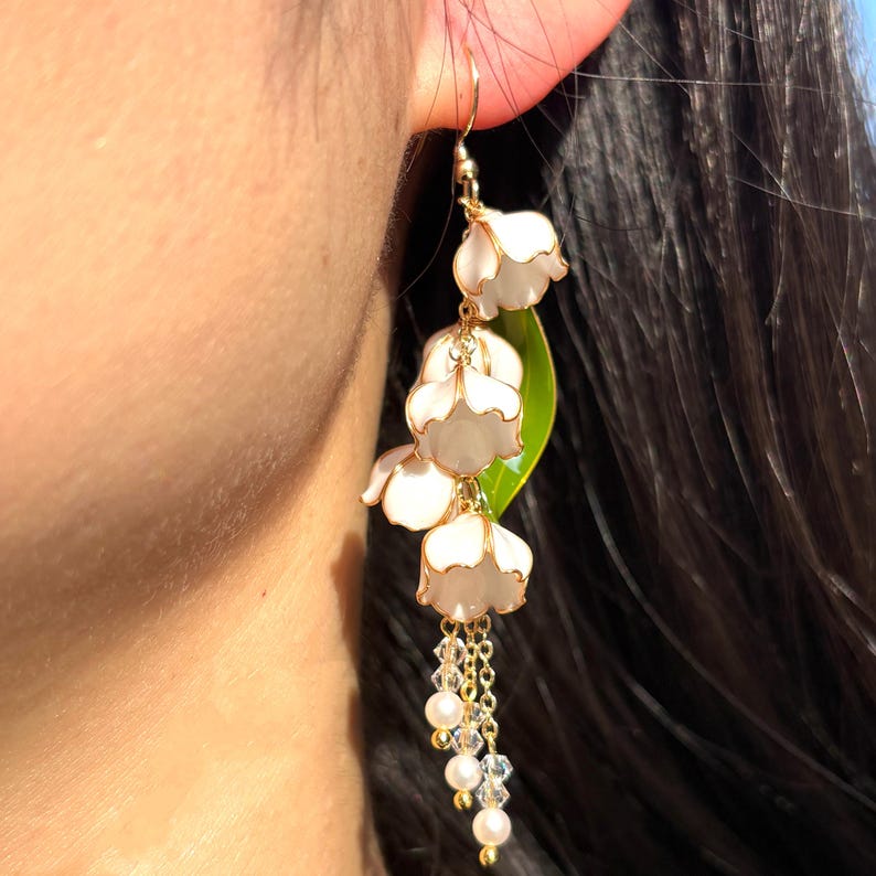 Op de afbeelding: Een bungelende oorbel met witte bloemhangers, goudkleurige accenten en een ketting van parels en kristallen. De oorbel bevat een groen blad. Een bloemvormige oorbel om in het oor te dragen.