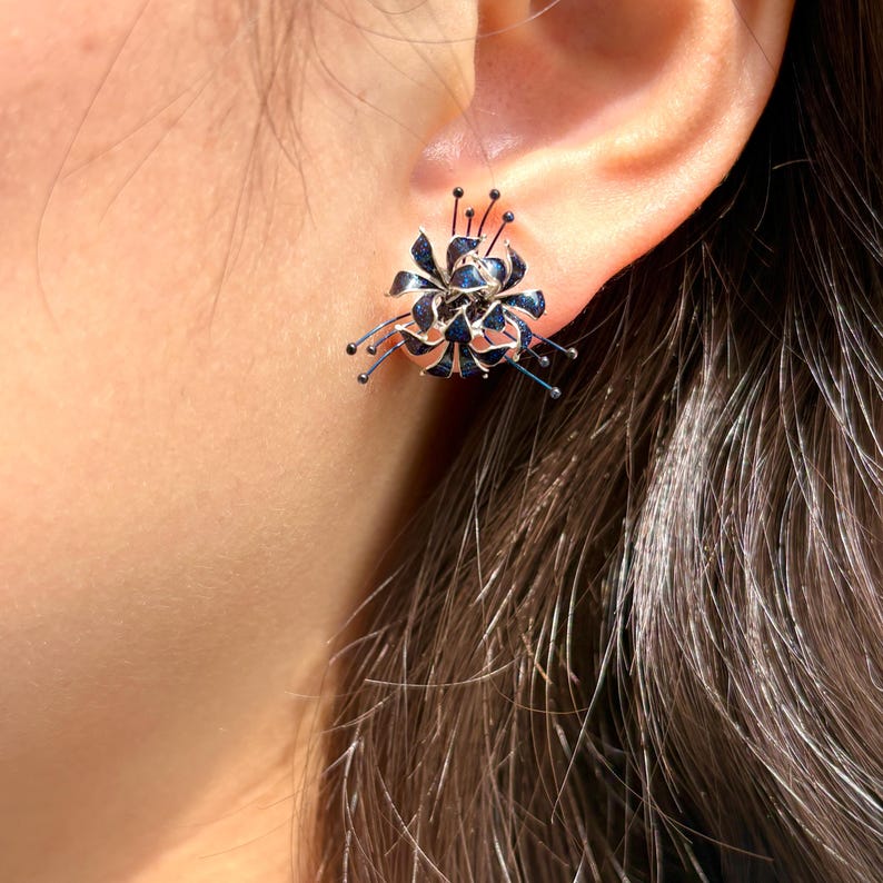 K&ouml;nnte beinhalten: Nahaufnahme eines bl&uuml;tenf&ouml;rmigen Ohrrings. Der Ohrring hat ein dunkelblaues Blumendesign mit silbernen Akzenten und kleinen schwarzen Perlen. Der Ohrring wird am Ohr einer Person getragen.